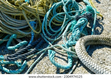 Heavy duty braided industrial fishing rope coiled. A large thick worn and woven style rope is piled next to a small frayed green cable fishing rope. The fishing gear is made of synthetic nylon cord.