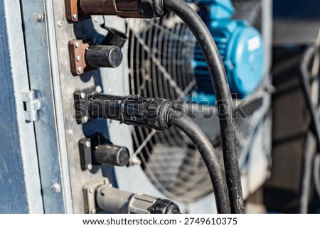 Heavy duty electrical cables and locking connectors plugged into outdoor power distribution panel, thick cords routed to equipment with cooling fan behind, industrial service hookup concept