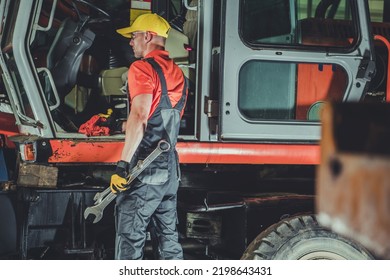 Heavy Duty Machinery Mechanic Standing In Front Of Orange Excavator With Large Wrenches In His Hands. Service And Repair Workshop.