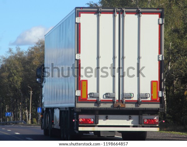 Heavy duty transport in logistics -\
white semi-trailer truck rides on asphalt highway in summer against\
green trees, road signs and blue sky, rear-side\
view