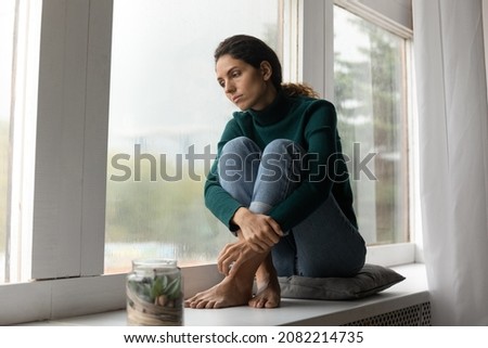 Heavy heart. Sad hispanic lady sit barefoot on windowsill feel absent minded tired stressed with problems in relations. Depressed young female look down the window try to overcome psychological trauma