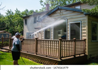 Heavy Husband Power Washing Large Windows On The Back Of A House Over A Brown Wooden Deck With A Pressure Washer