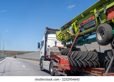 Heavy industrial truck with low side on low-frame platform transports disassembled big green combine harvester along ordinary highway on summer day. - Shutterstock ID 2232954643