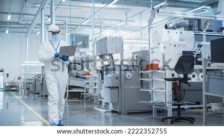 Heavy Industry Manufacturing Factory: Scientist in Sterile Coverall Walking with Laptop Computer, Examining Industrial CNC Machine Settings and Configuring Production Functionality.