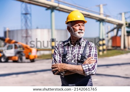 Heavy industry, power plant, oil refinery, and factory staff. An experienced senior factory worker with a helmet on his head standing in a refinery yard with arms crossed and looking away.