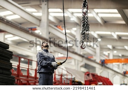 A heavy industry worker relocates chains on an industrial hook by using a controller while standing in the factory.