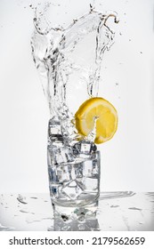 Heavy And Large Splash Of An Ice Cube In A Glass Of Fizzy Drink Decorated With A Lemon. Splash On White Background With Reflection On The Base.