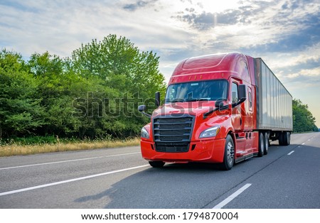 Heavy loaded classic red big rig semi truck with high roof transporting commercial cargo at dry van semi trailer running on the straight wide divided multiline highway road for timely delivery