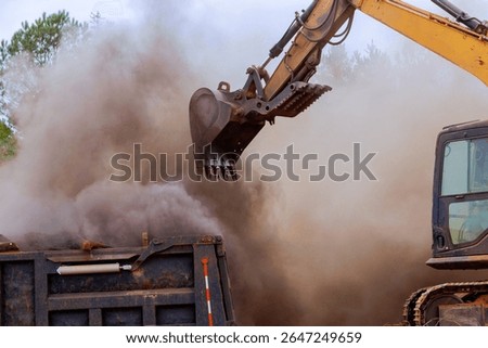 Heavy machinery operates to load construction debris into truck, creating cloud of dust at works site.
