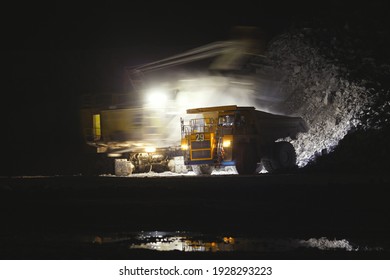 Heavy mining dump truck during a night loading of rock in a limestone quarry, stands on the background of unsharp mine excavator which is in motion, shot at slow shutter speeds.  Mining industry.