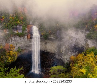 A heavy morning fog hangs over a waterfall surrounded by autumn foliage as it cascades over a rocky cliff into a dark pool at Fall Creek Falls State Park, Tennessee. - Powered by Shutterstock