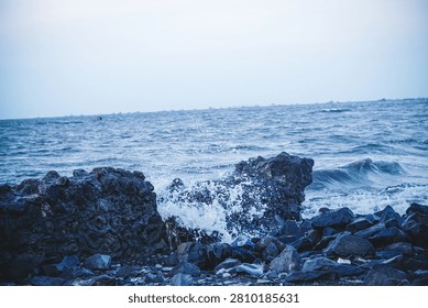 Heavy ocean waves break and splash over a rough stone barrier on the coastline of Tanjung Pasir, Indonesia, under a dim late afternoon sky.