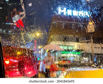 Heavy rain in the night, rain on car window in focus, through the glass our of focus we can see a woman holding an umbrella and shopping bags, street traffic and streetlights, with bright colours