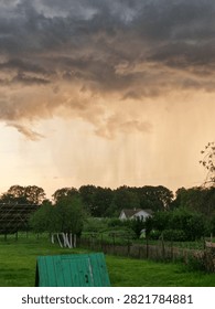 Heavy rain wall pouring from dramatic storm clouds over a rural village at sunset. Countryside house, green fields, extreme weather, downpour and climate change concept.