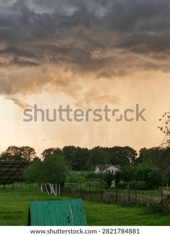 Heavy rain wall pouring from dramatic storm clouds over a rural village at sunset. Countryside house, green fields, extreme weather, downpour and climate change concept.