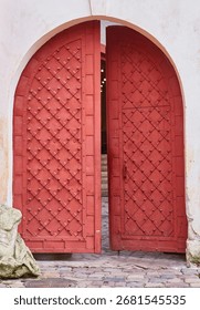 A heavy, rustic red medieval-style gate in a stone archway stands slightly ajar. The ancient door is decorated with a diamond pattern and metal studs.