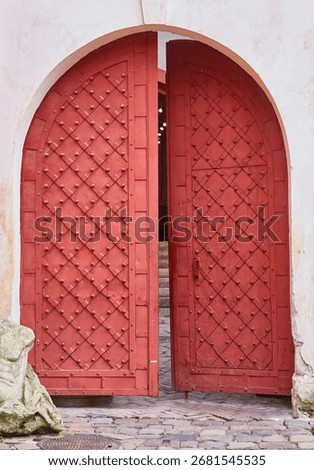 A heavy, rustic red medieval-style gate in a stone archway stands slightly ajar. The ancient door is decorated with a diamond pattern and metal studs.