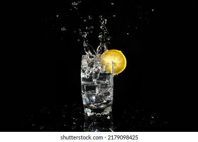 Heavy Splashing Of An Ice Cube In A Glass Of Fizzy Drink Decorated With A Lemon. Splash On Black Background With Reflection On The Base. Vertical Format. Hard Ligth