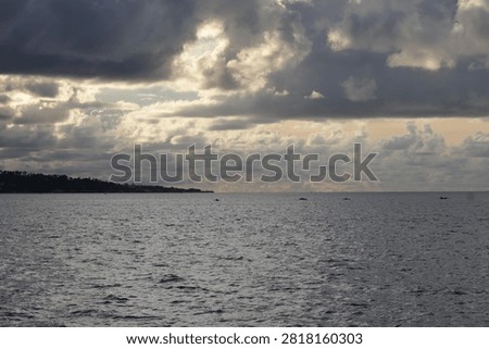 Heavy storm clouds break with soft light over a calm coastal sea in Indonesia, silhouetting a tree-lined coastline and small fishing boats scattered across the water in a moody, atmospheric seascape.