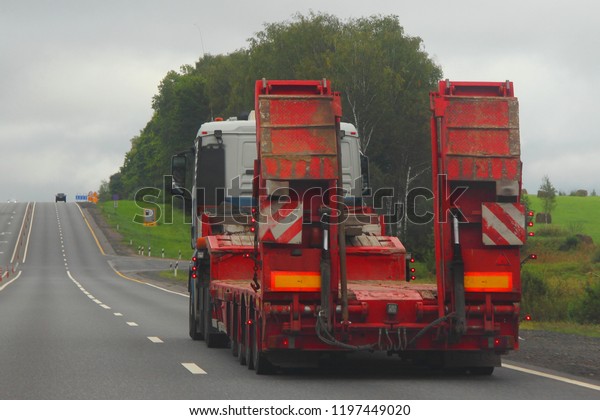 Heavy truck with red\
low-frame four-axle semi-trailer trawl on a two-lane asphalt\
highway road in the summer day, rear-side view – Logistics,\
transportation, trucking\
industry