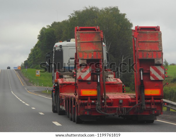 Heavy truck with red\
low-frame four-axle semi-trailer trawl on a two-lane asphalt\
highway road in the summer day, rear-side view – Logistics,\
transportation, trucking\
industry