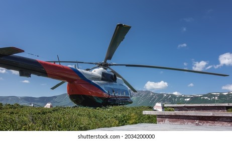 The helicopter is standing on a green meadow in the caldera of an extinct volcano. A picturesque mountain range against the blue sky. In the foreground are the steps of a wooden staircase. Kamchatka