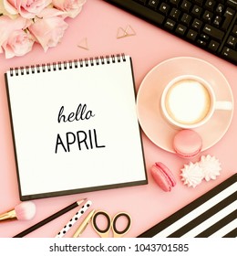 Hello April text on notebook. Office desk table with cup of coffee, sweet snacks, computer and pink flowers. Pink, gold, black and white tones. Top view, flat lay.