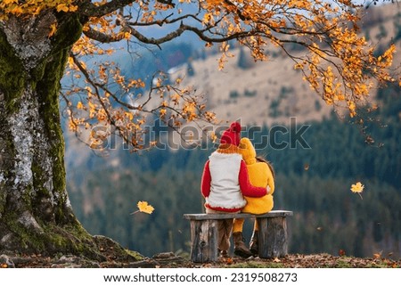 Hello autumn. Children are sitting on a wooden bench under a old tree with autumn leaves. Family walk in autumn.