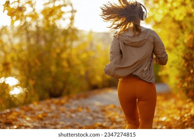 Hello autumn. Seen from behind woman in fitness clothes in the park with headphones running.
