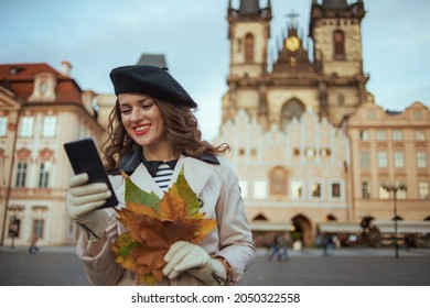 Hello autumn. smiling 40 years old woman in beige trench coat and black beret with autumn yellow leaves using smartphone applications at Old Town Square in Prague.