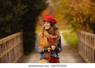 Hello autumn. smiling elegant woman in jeans shirt and red hat with autumn leaf, scarf and gloves in the city park.