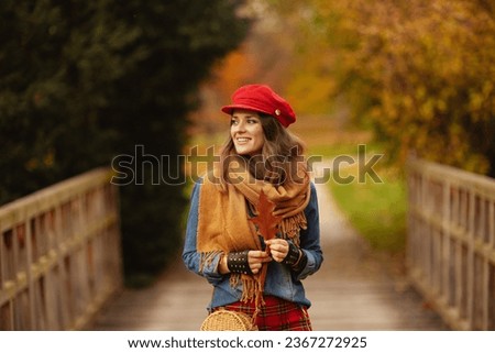 Hello autumn. smiling elegant woman in jeans shirt and red hat with autumn leaf, scarf and gloves in the city park.