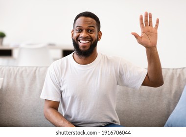 Hello. Cheerful African American Man Waving Hand Smiling To Camera Sitting On Couch At Home. Hi Greeting Gesture Concept. Video Call And Modern Remote Communication