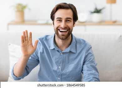 Hello. Happy Man Waving Hand Smiling To Camera Sitting On Couch At Home. Cheerful Guy Gesturing Hi Greeting Making Video Call Indoors. Modern Remote Communication Concept