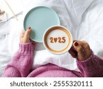 Hello new year 2025 celebrated coffee cup with number 2025 over frothy surface of cappuccino holding by woman in pink knitted sweater sitting on the bed with white blanket, notebook and pencil.