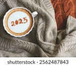 Hello new year 2025 celebrated coffee cup with number 2025 over frothy surface of cappuccino served in white coffee mug on soft brown knitted sweater background. Holidays food art theme.