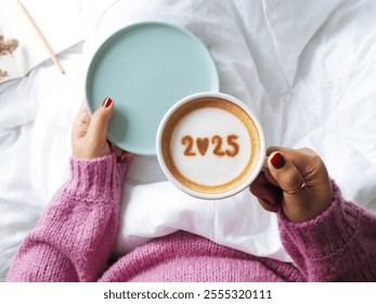 Hello new year 2025 celebrated coffee cup with number 2025 over frothy surface of cappuccino holding by woman in pink knitted sweater sitting on the bed with white blanket, notebook and pencil. - Powered by Shutterstock