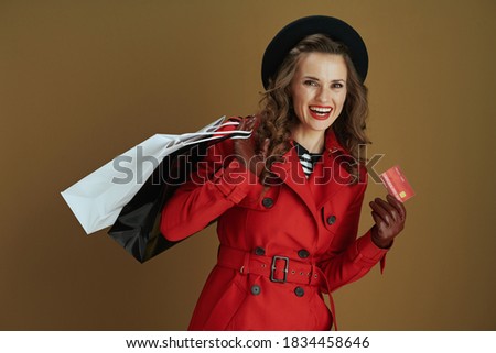 Hello october. smiling modern middle aged woman in red coat and black beret with credit card, leather gloves and paper shopping bags against bronze background.