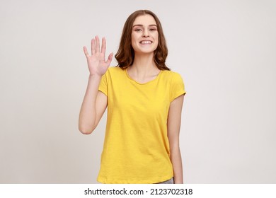 Hello! Portrait of charming teenager girl with brown hair, wearing casual style outfit standing, waving hand and saying Hi to camera with toothy smile. Indoor studio shot isolated on gray background. - Shutterstock ID 2123702318