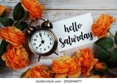 Hello Saturday Card And Alarm Clock With Orange Flower Decoration On Wooden Background