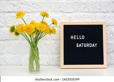 Hello Saturday words on black letter board and bouquet of yellow dandelions flowers on table against white brick wall. Concept Happy Saturday. Template for postcard.