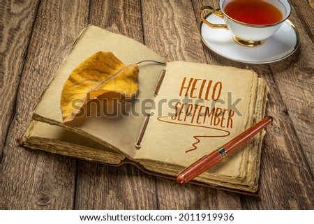 Hello September handwriting in a retro journal with decked edge handmade paper pages and a stylish pen on a rustic wooden table with a cup of tea, fall and journaling concept