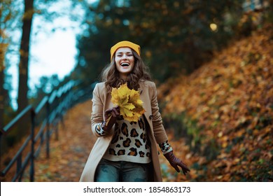 Hello september. happy trendy female in brown coat and yellow hat with autumn yellow leaves enjoying promenade outside in the city park in autumn.