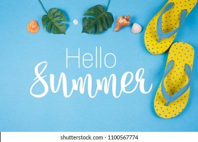Hello Summer text on blue background, Yellow sandals with green tropical palm leaf and shell. Beach accessories