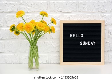 Hello Sunday words on black letter board and bouquet of yellow dandelions flowers on table against white brick wall. Concept Happy Sunday. Template for postcard.