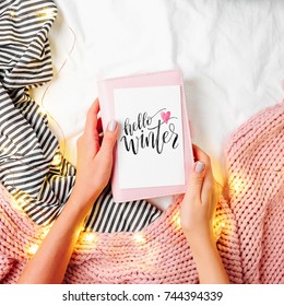 Hello winter! Female hands hold postcard. Bedding sheets with fairy lights. Copy space. Flat lay, top view