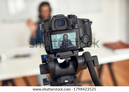 Hello. Young happy male teacher looking at camera and waving while giving online class through webcam at home. Tutor recording video tutorial. Focus on a camera screen. E-learning. Distance education