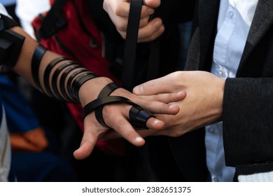 With the help of another adult, a bar mitzvah boy wraps the black leather straps of his tefillin around his arm, hand and fingers in preparation for Jewish morning prayers. 