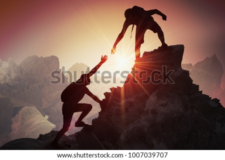 Help and assistance concept. Silhouettes of two people climbing on mountain and helping.