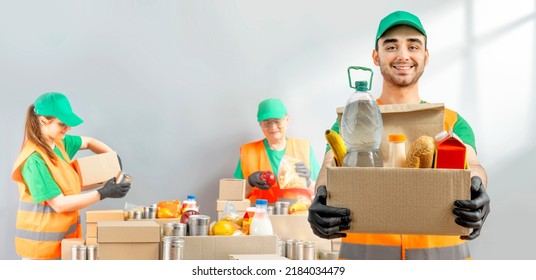 Help Collection Center, Free Food Distribution. Volunteer Carrying Food Donation Box. Young Smiling Man Wearing Green Uniform Cap And T-shirt, Orange Vest Holds Out Grocery Set For In-need People.